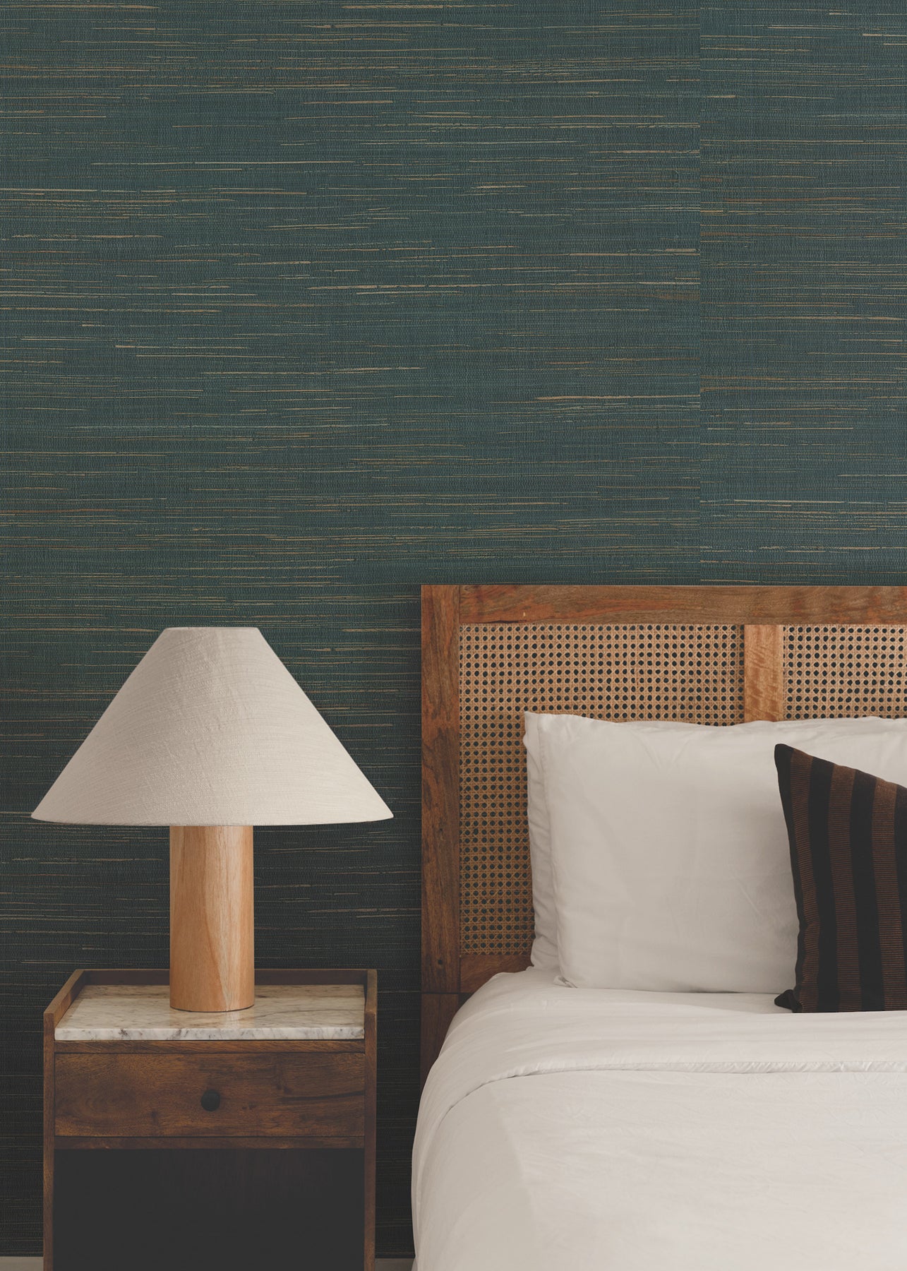 Bedroom with wooden bed and cane headboard, white bedding, striped pillow, wooden nightstand, beige lamp, teal textured wall.