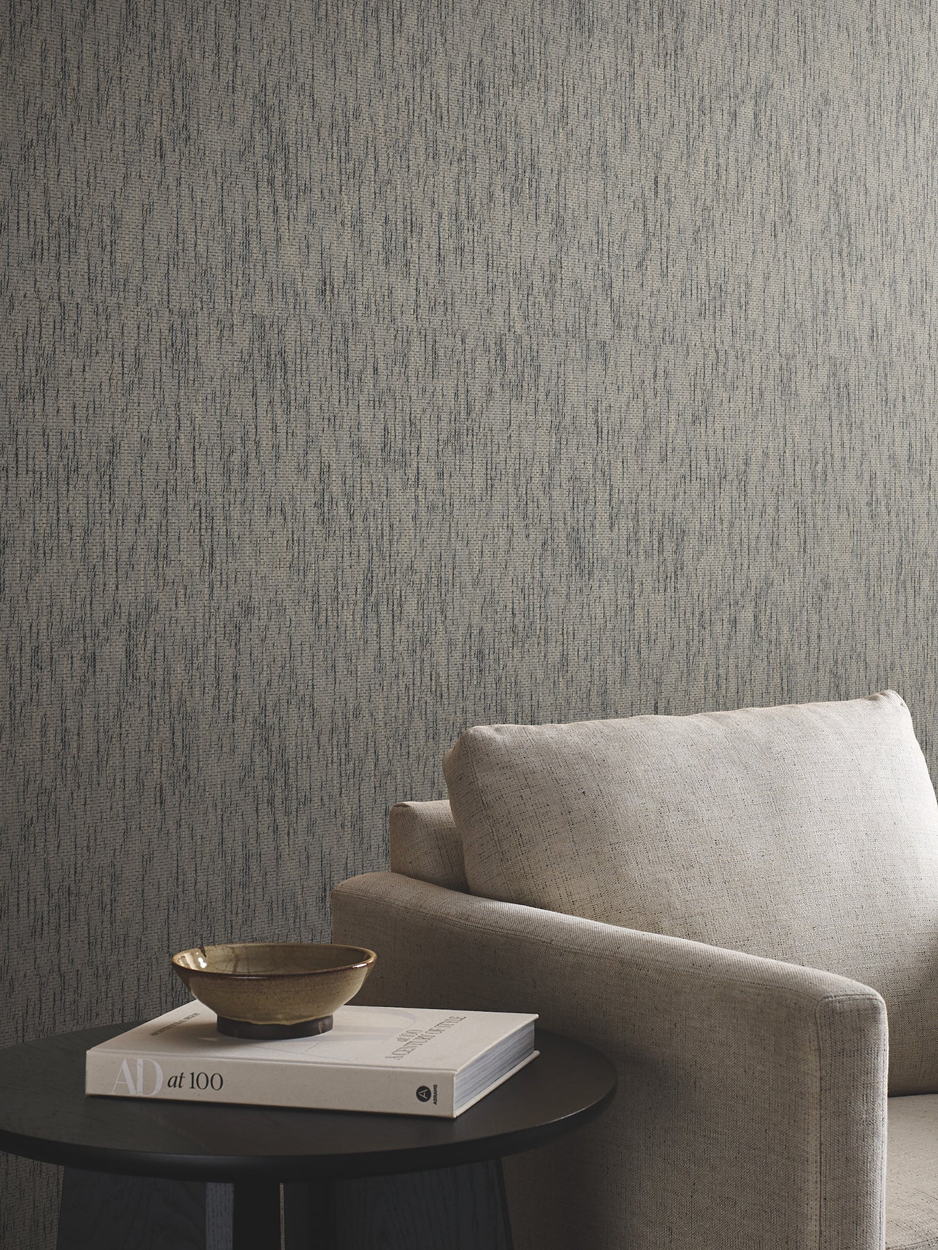 Beige fabric sofa with cushion beside a round black side table holding a beige bowl and stacked books, grey textured wall.
