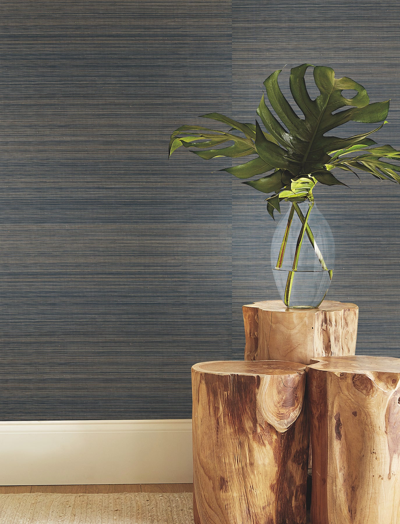 Blue ombre grasscloth wallpaper backdrop with a clear glass vase, monstera leaves, and stacked natural wood stumps in the foreground.
