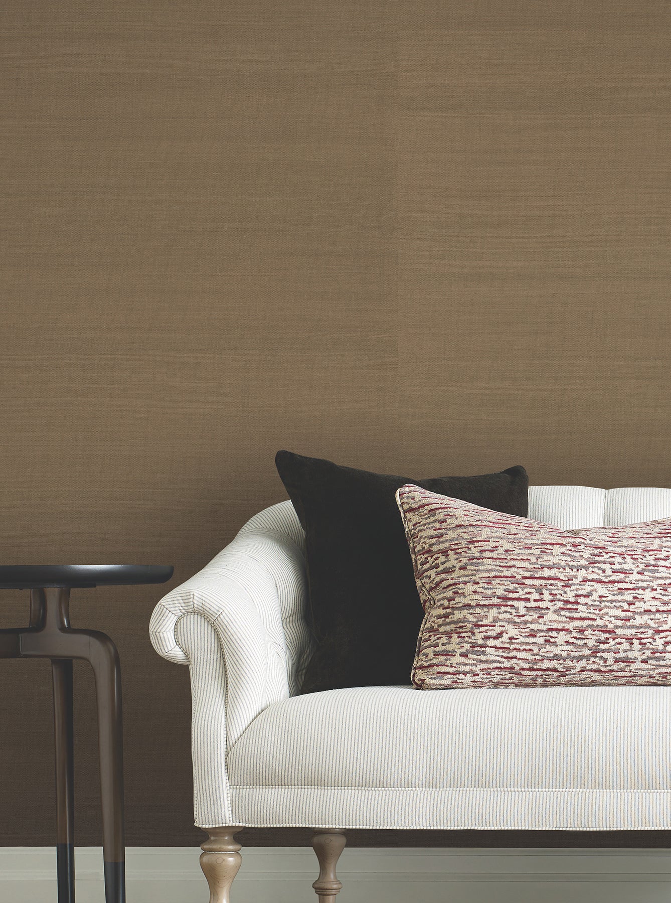 Room showing brown toffee grasscloth wallpaper on the walls behind a white rolled-arm sofa with black and red-beige patterned pillows and a dark wood side table.