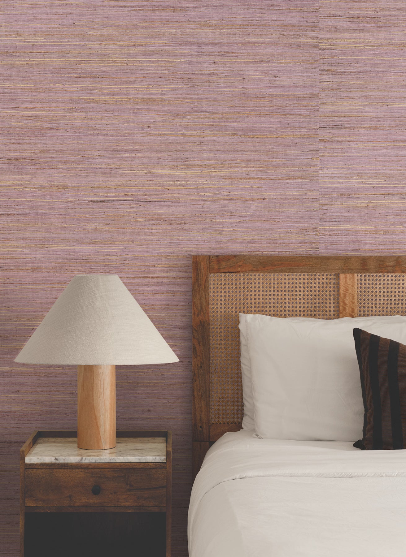 Bedroom with pink striped wallpaper, wicker headboard, white bedding, dark accent pillow, wooden nightstand, beige lamp.
