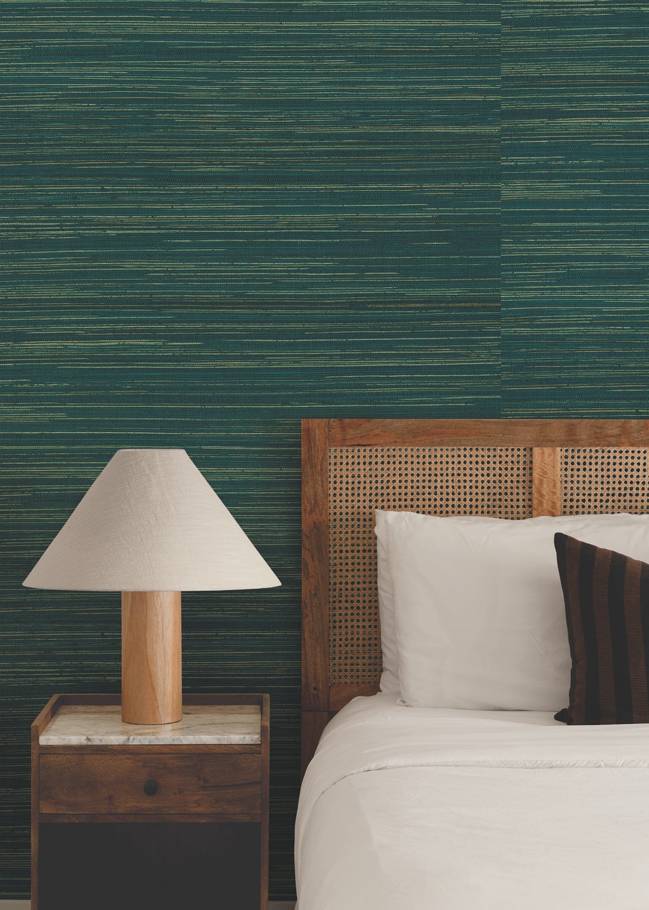 Bedroom with a cane-panel wooden headboard, white linens, a dark throw pillow, wooden nightstand, and beige lampshade against teal striped wallpaper.