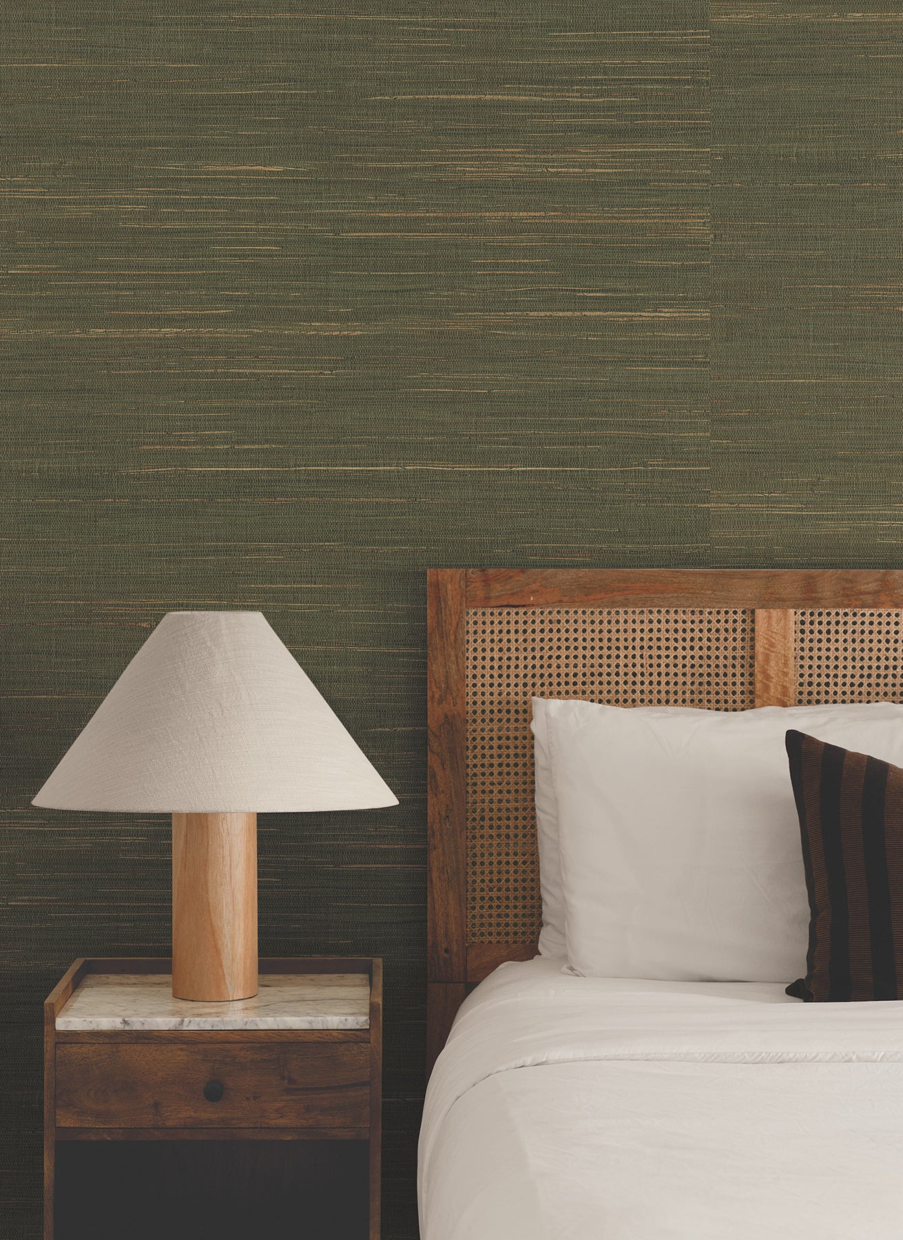 Wooden bed with cane headboard, white linens, dark striped pillow; bedside lamp on a wooden nightstand against green wall.