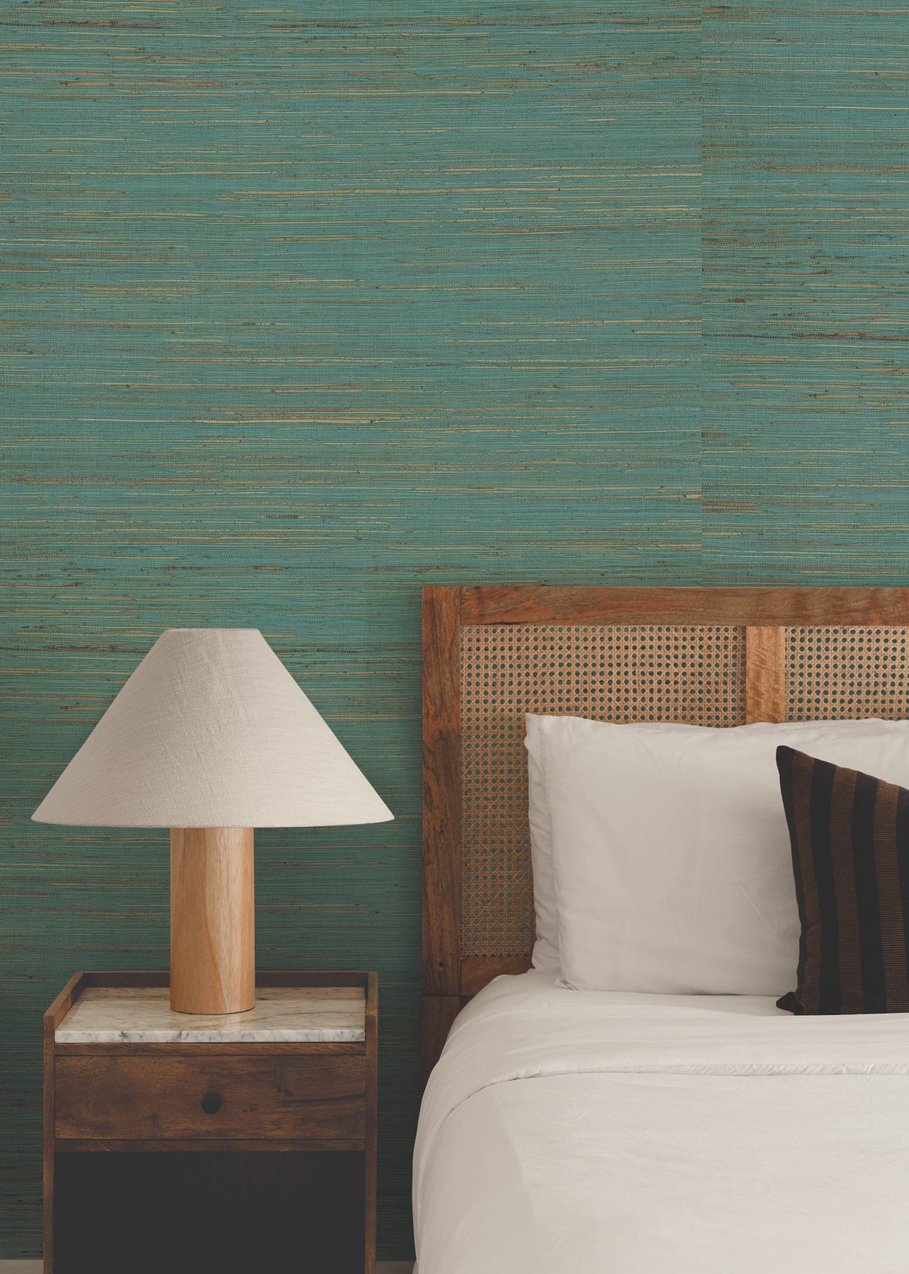 Bedroom with teal distressed wall, wooden headboard, white bedding, striped pillow, and nightstand with beige lampshade.