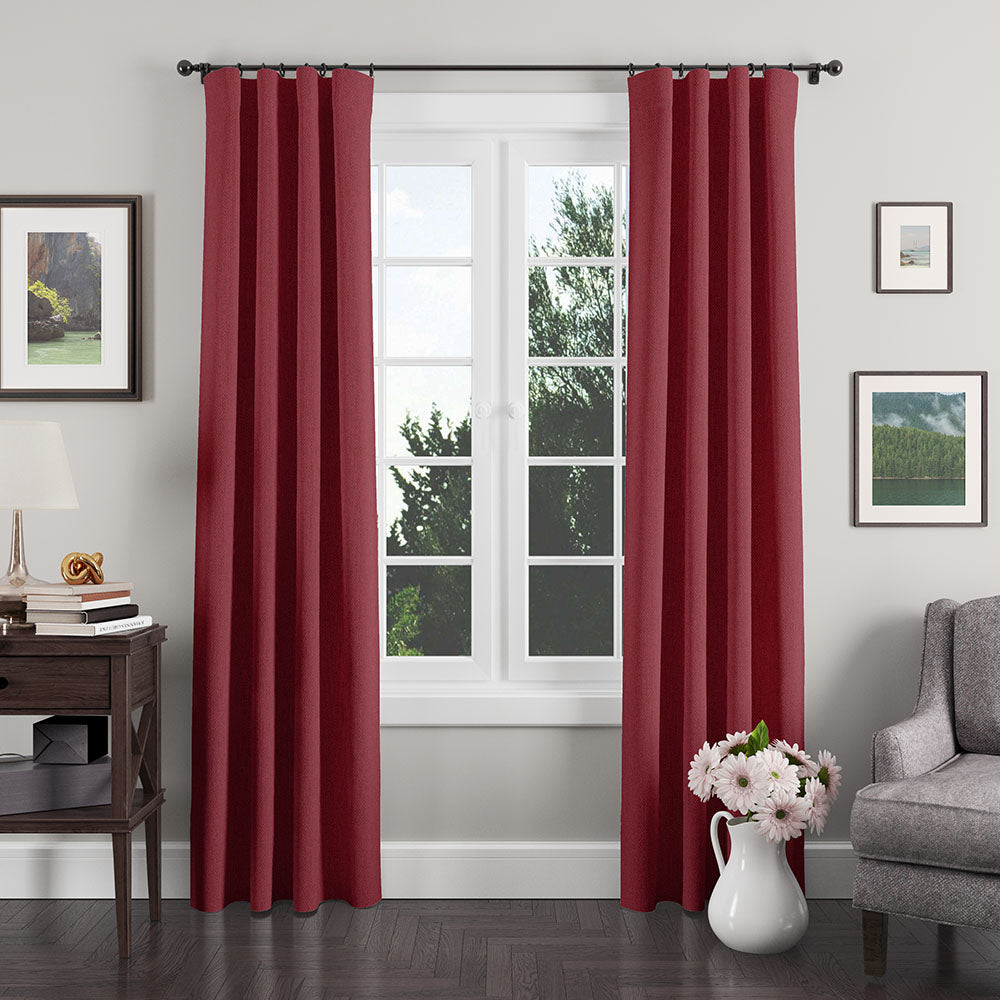 Deep red curtains on a black rod framing a window in a cozy living room with a gray sofa and white vase of pink flowers.