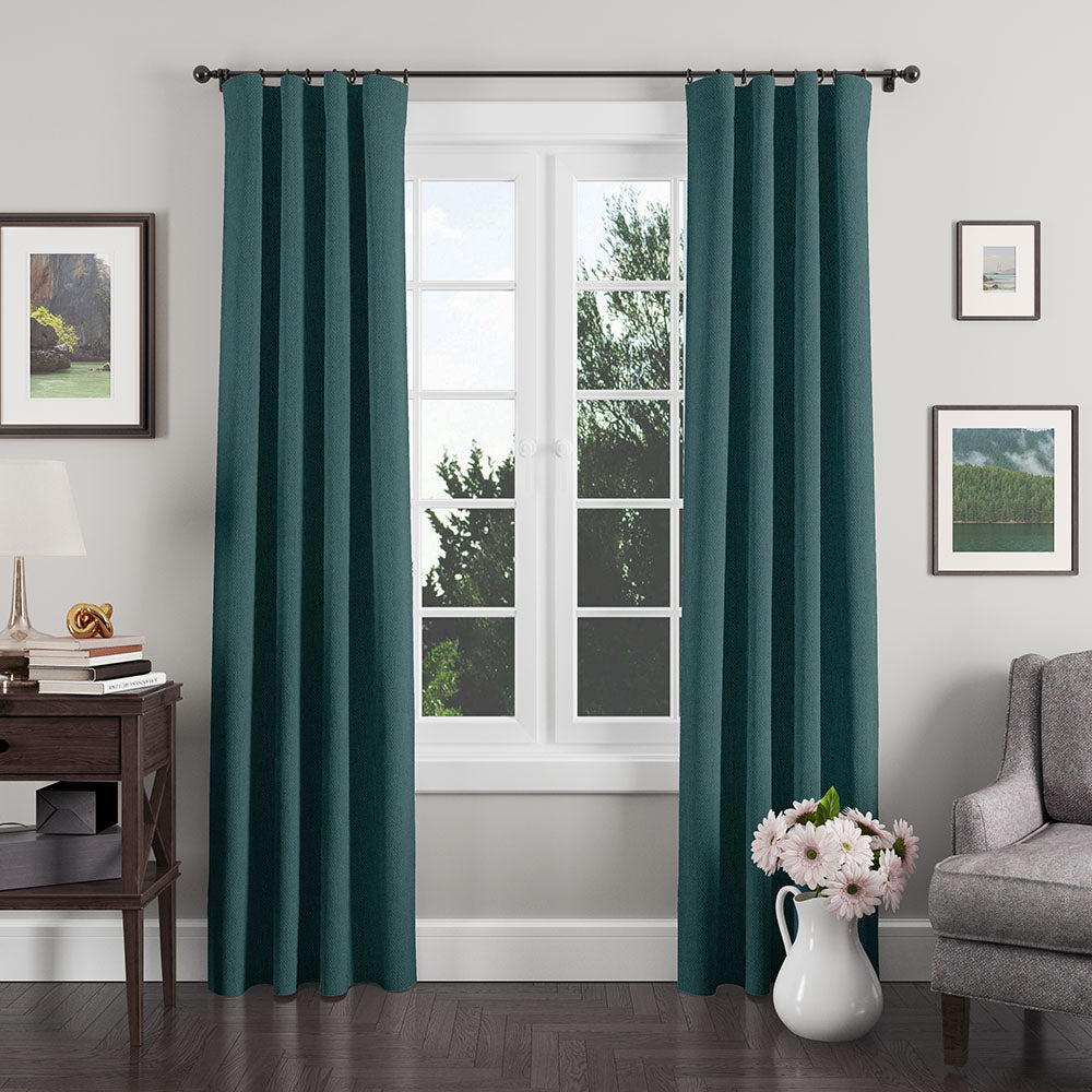 Teal curtains frame a white-framed window in a living room with a side table, lamp, wall art, gray sofa, and pink flowers.