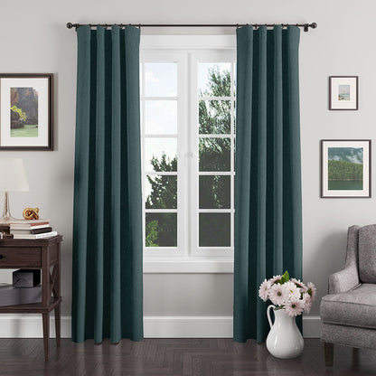 Teal curtains frame a white window in a cozy living room; gray armchair, dark wood side table, lamp, & vase of pink flowers.