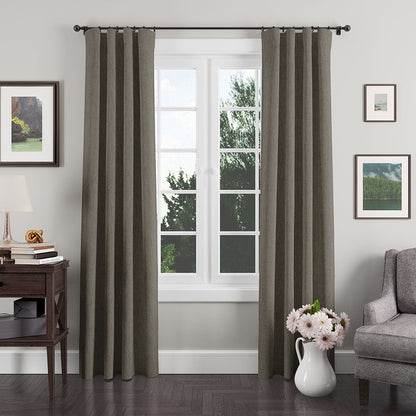 Olive-gray textured curtains frame a white mullioned window; dark wood console, lamp, art, gray sofa, and pink flower vase.