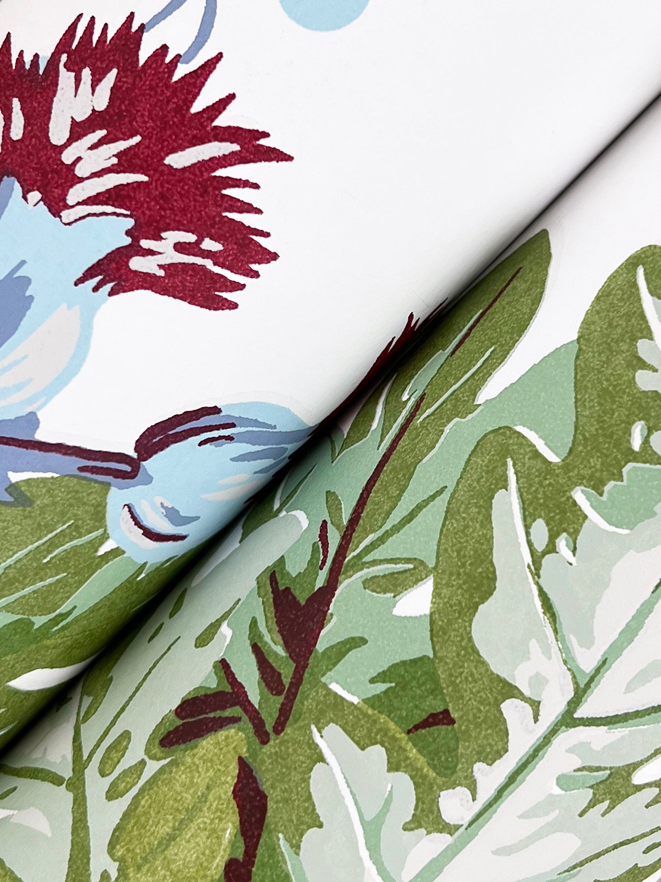 Folded wallpaper sample showing a botanical leaf pattern in greens on a white background with subtle maroon accents and pale blue highlights.