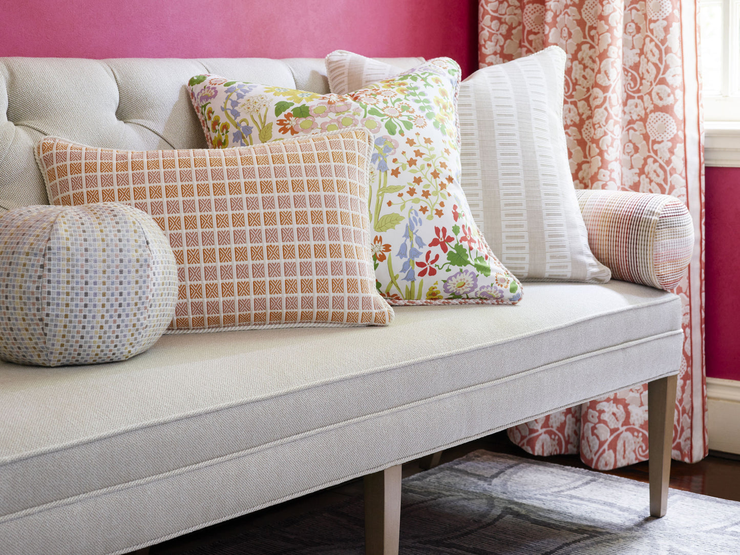 Beige fabric sofa with tufted back and wooden legs, colorful throw pillows (plaid, floral, striped) against a pink wall.