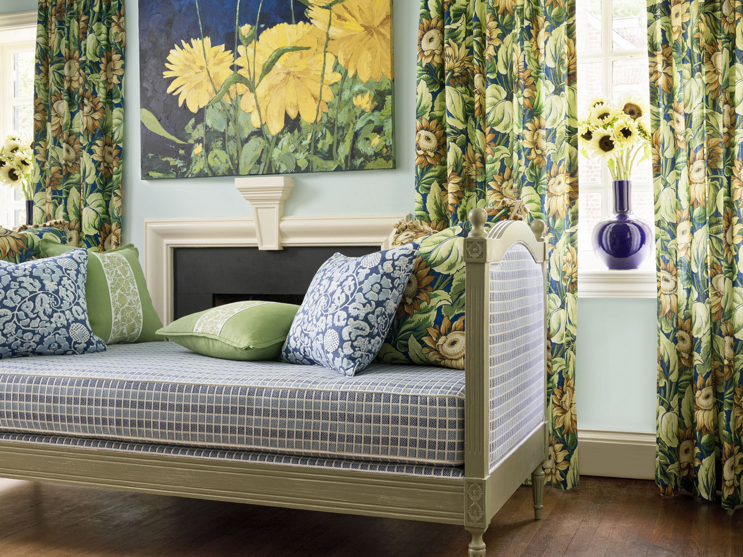 Beige daybed with denim floral fabric upholstery, blue-check mattress, green and blue cushions, tropical floral curtains and yellow flower artwork.