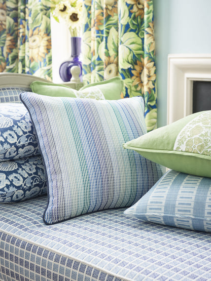 Blue and denim Maiden Floral fabric throw pillow on a bed with layered blue pillows; botanical curtains and a purple vase.