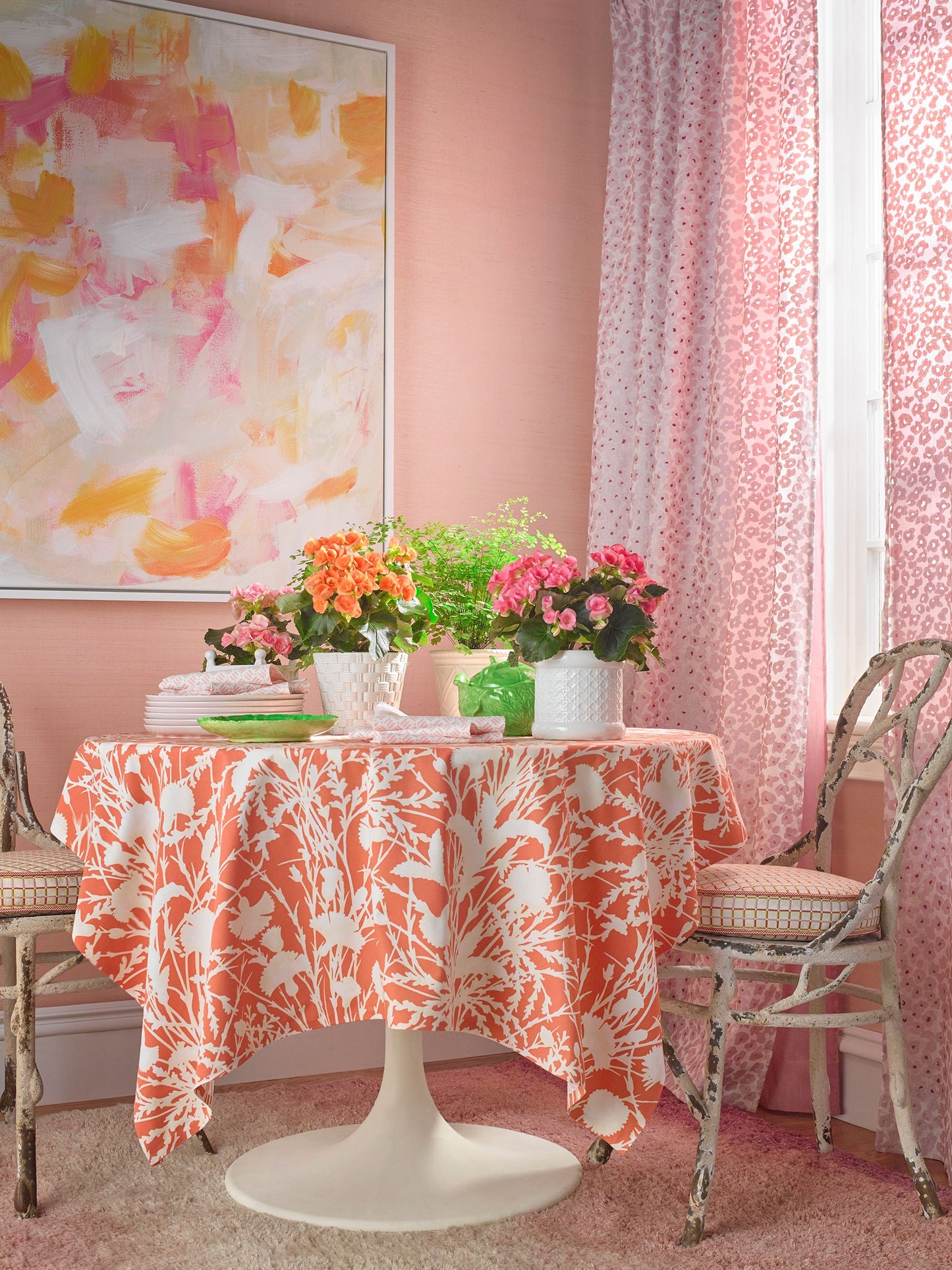 Round pedestal table with orange botanical tablecloth, potted pink and orange flowers, wicker chairs, pink walls.