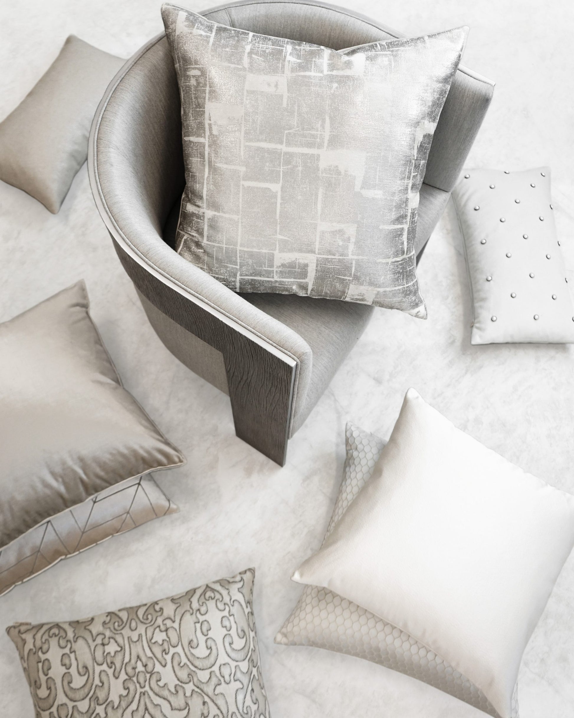 Grey curved armchair with a metallic-patterned silver cushion; surrounded by beige and silver throw pillows on a light floor.