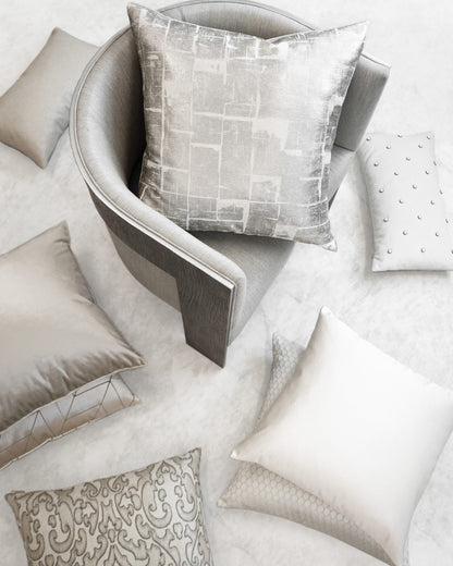 Grey curved armchair with a metallic-patterned silver cushion; surrounded by beige and silver throw pillows on a light floor.