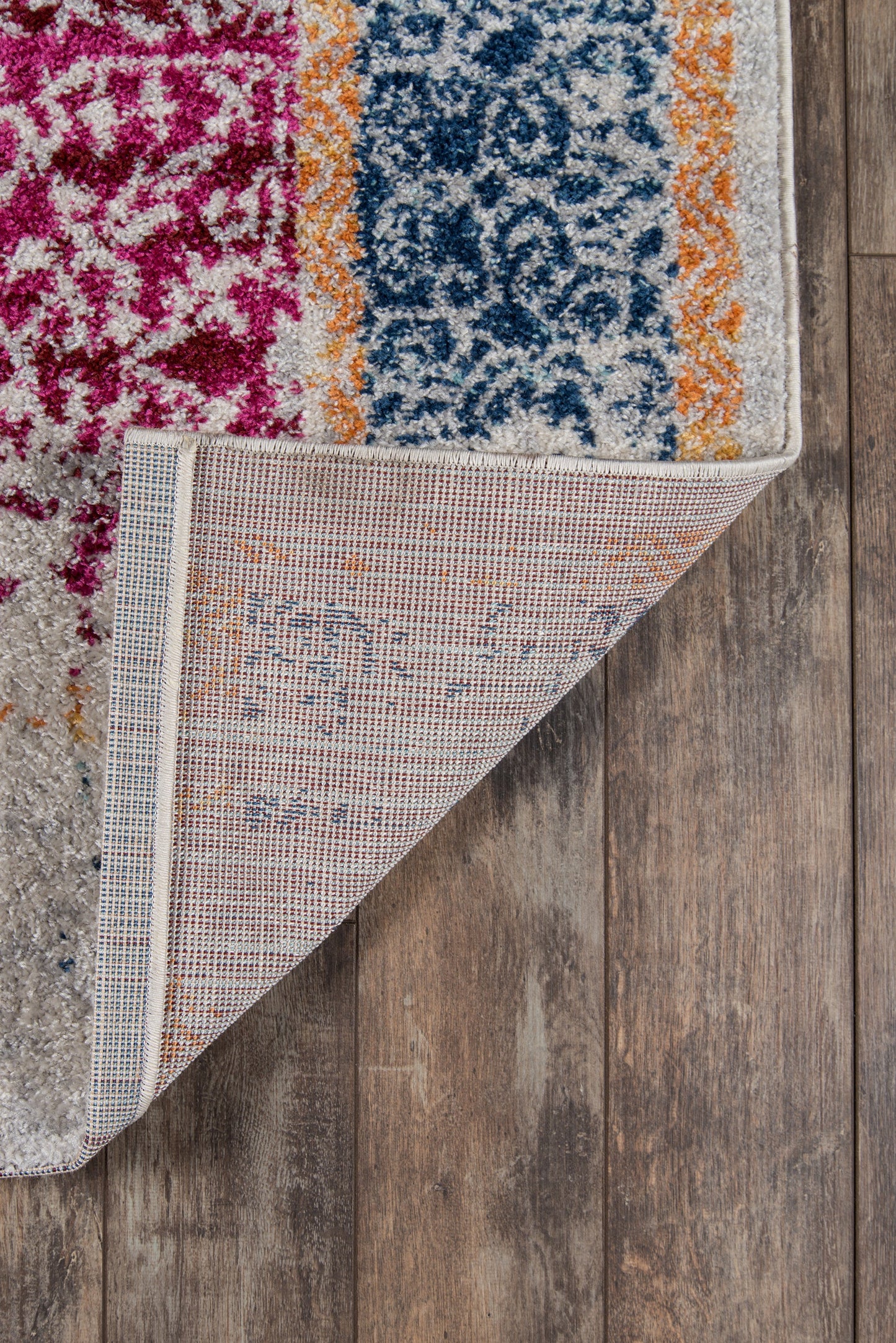 Vibrant patterned area rug with pink magenta, blue ornate field, orange border, folded corner reveals beige backing on wood floor.