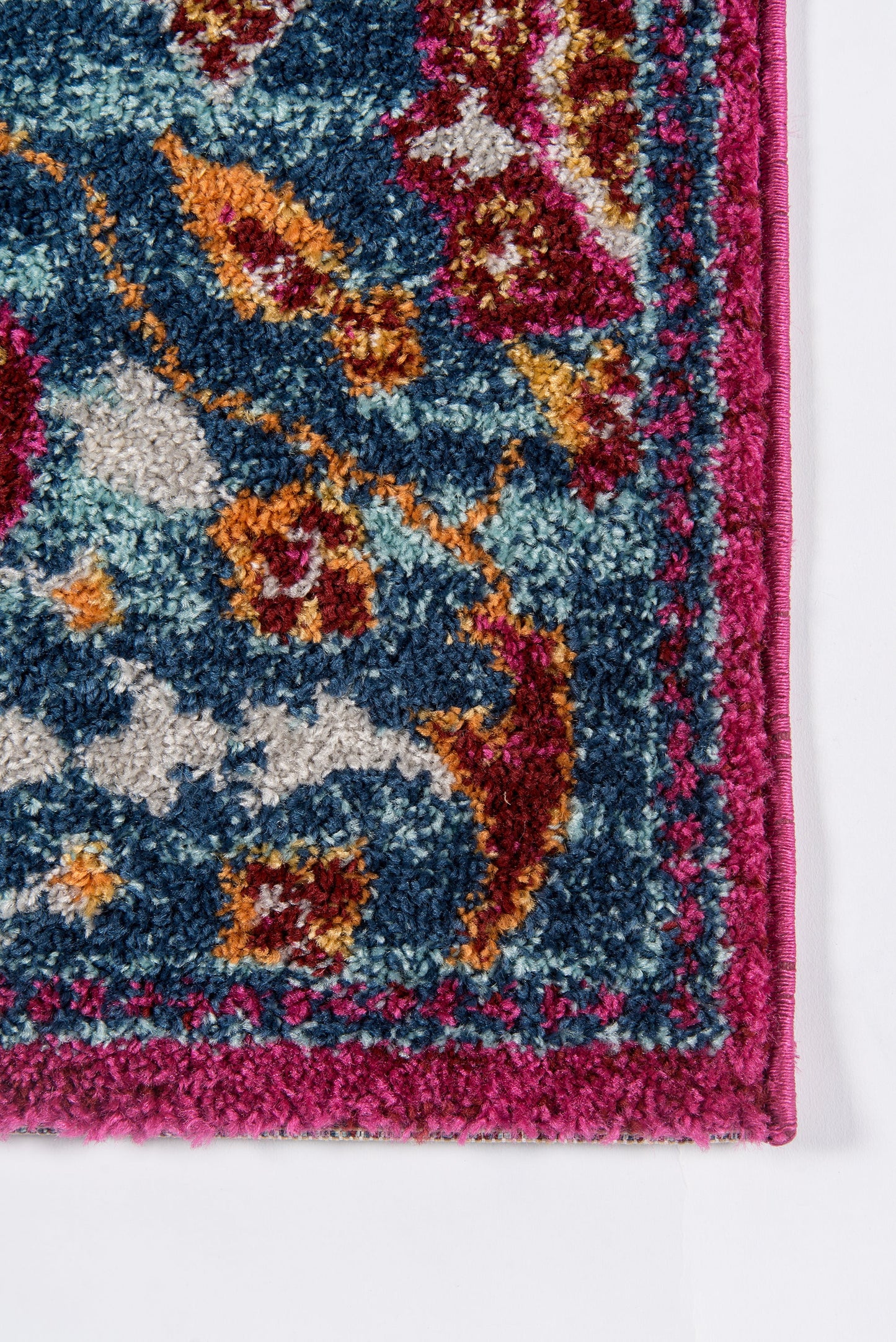 Close-up of a plush, vibrant patterned rug with blue field, magenta border and pink trim; floral motifs in teal, orange and gold.