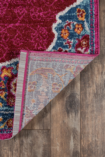 Vibrant pink rug with blue floral border and pink trim; folded corner reveals underside; on hardwood floor.