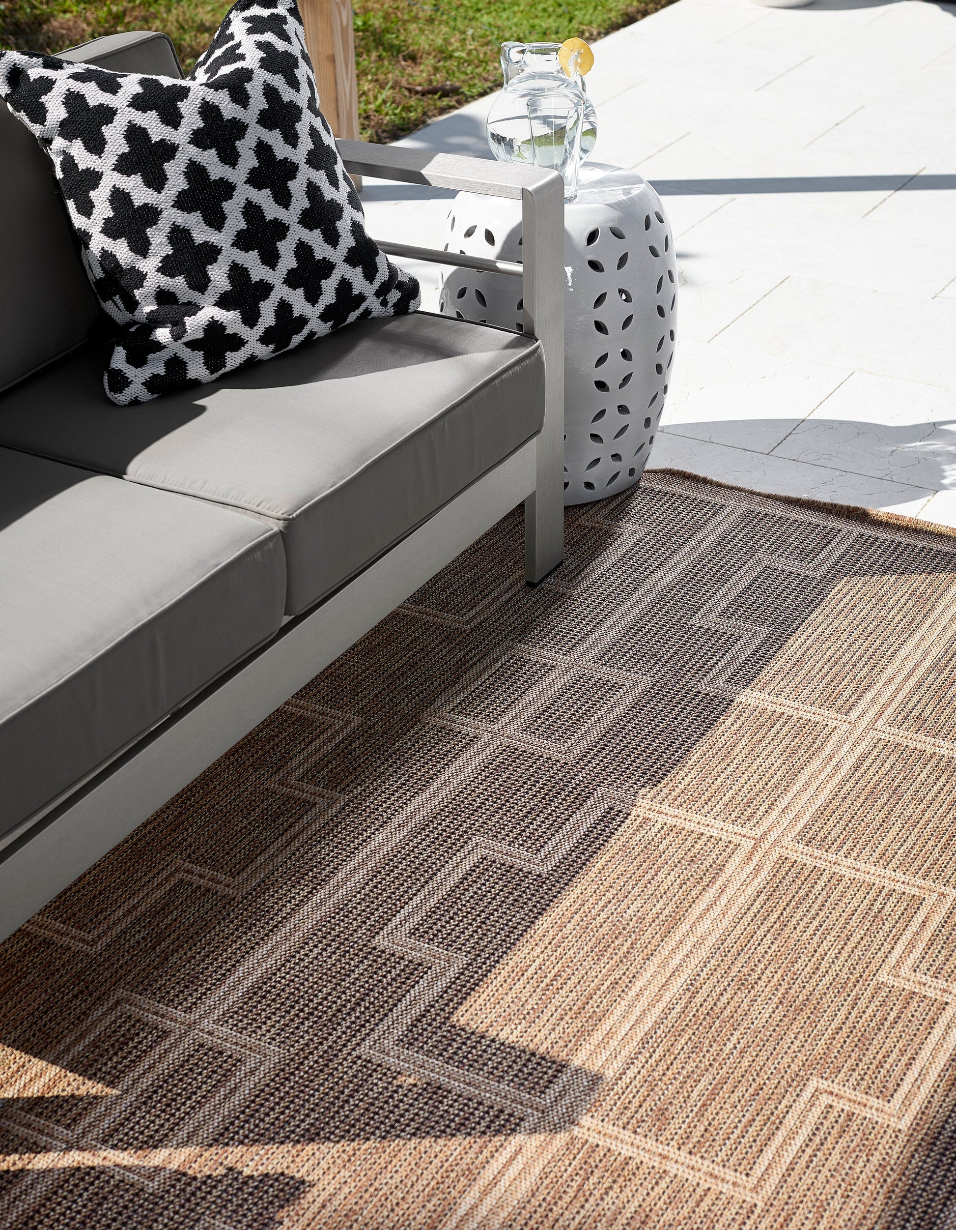 Beige geometric rug on outdoor patio; visible items include gray cushions, a white perforated side table, a black-and-white pillow and a pitcher.