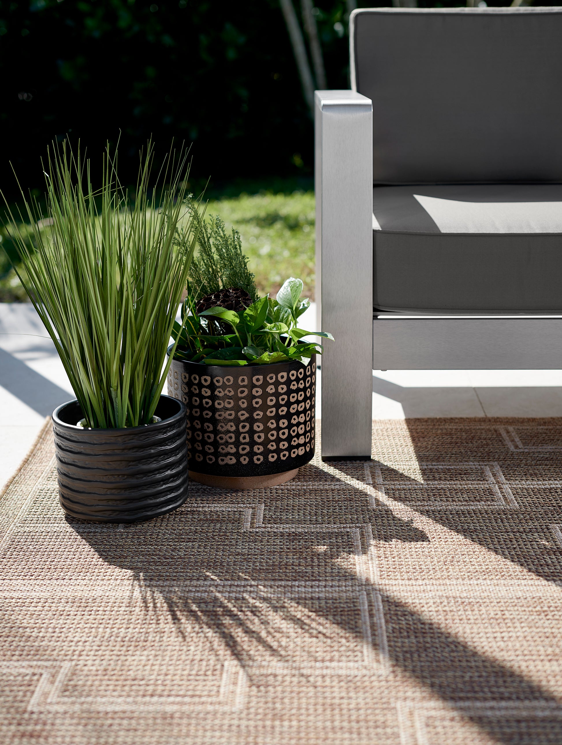 Outdoor patio: two black planters with tall grass and leafy greens beside a gray metal-frame chair on a beige woven rug.