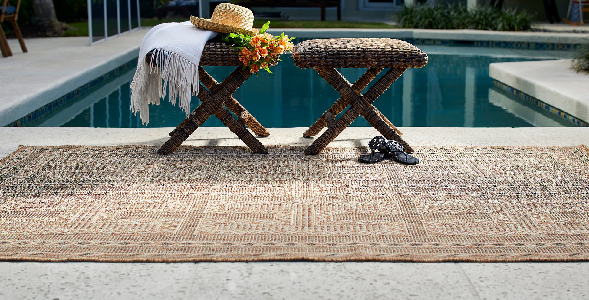 Beige woven outdoor rug by poolside with wicker ottomans, straw hat, orange flowers, white fringe throw; black sandals.