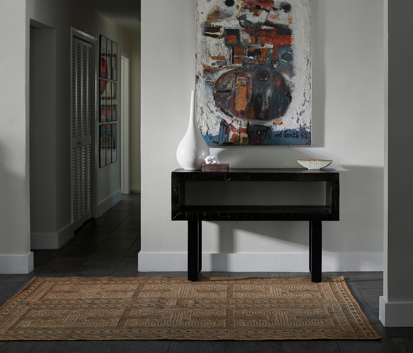 Dark wood console table with open lower shelf, white vase and small decor; abstract painting above, woven jute rug beneath.