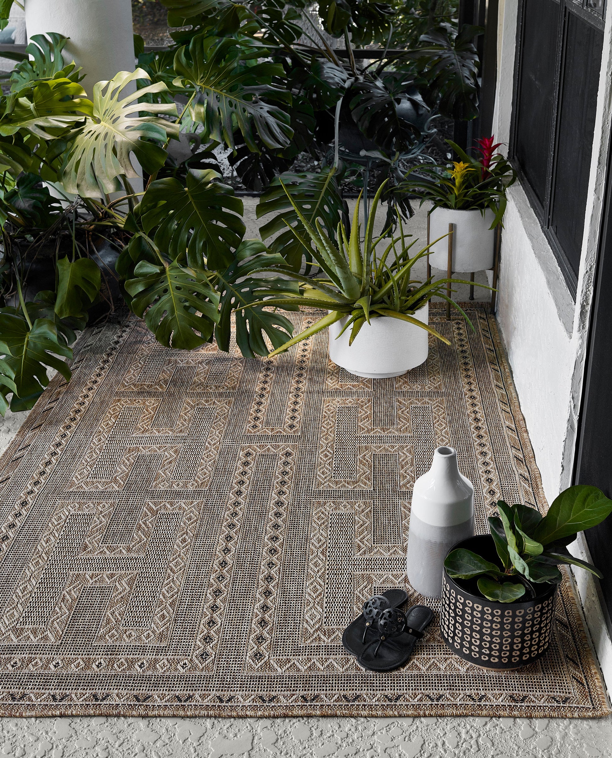 Boho porch with beige geometric rug, monstera plants in white pots, a gray vase, black sandals, and a dotted planter.