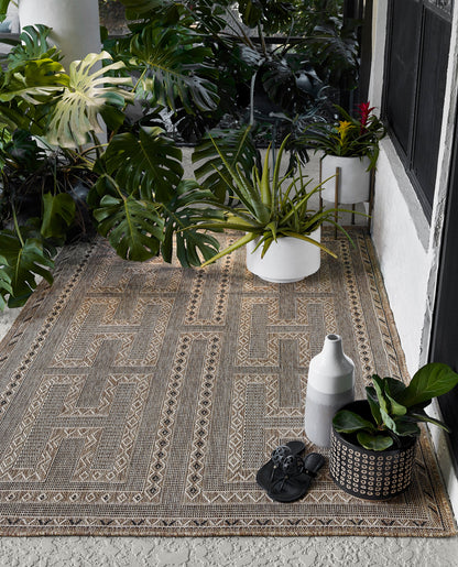 Beige outdoor rug with geometric border on concrete porch, framed by monstera leaves, aloe in white pots, a gray vase, and black sandals.