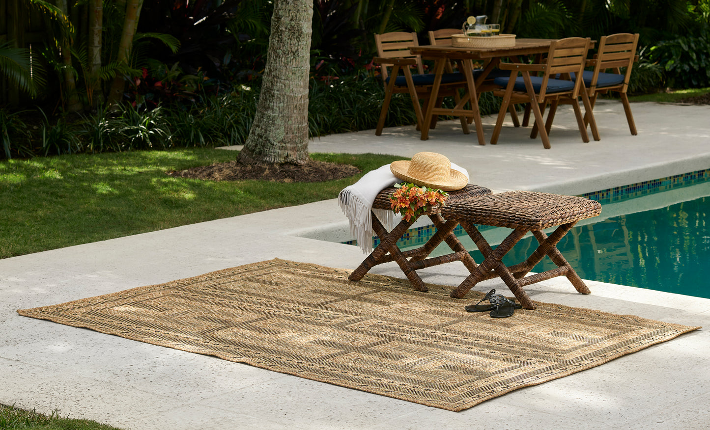 Natural jute rug with geometric pattern on poolside patio, woven wicker coffee table, hat, flowers, background dining set