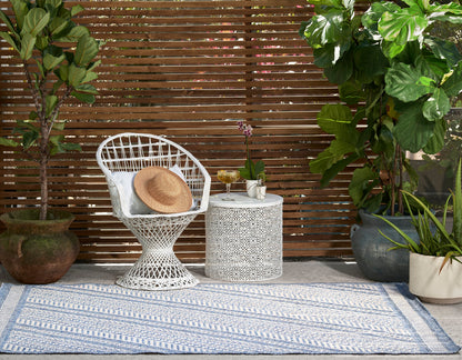 Boho outdoor scene featuring white wicker chair, straw hat, circular lattice table, blue patterned rug, & lush potted plants.