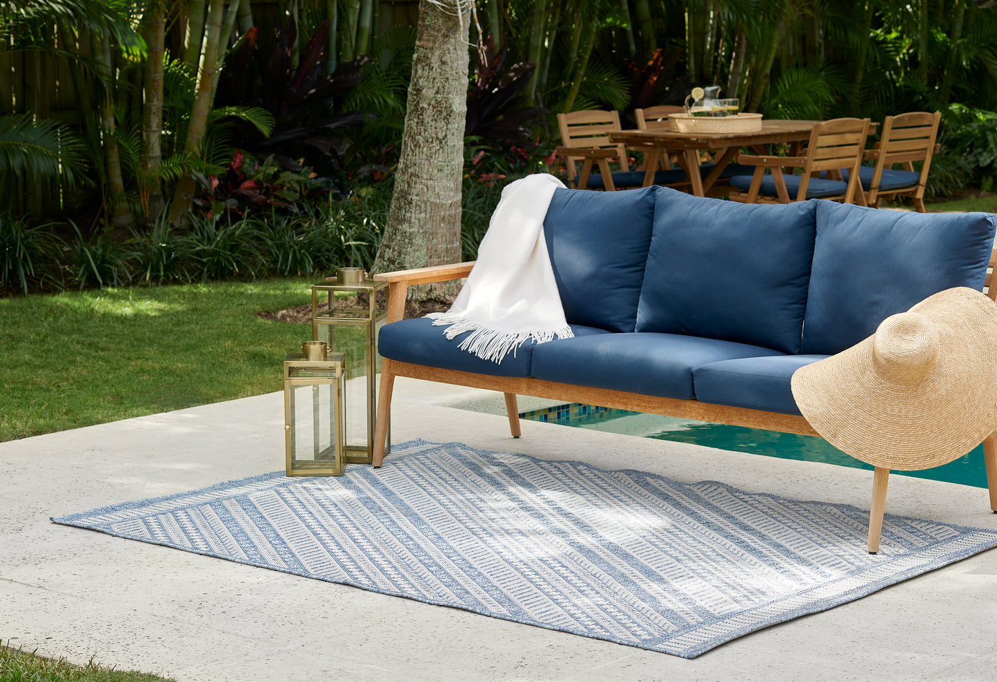 Outdoor patio with navy cushioned wooden sofa, white throw, striped blue rug; brass lanterns & wide-brim straw hat.