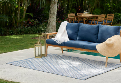 Outdoor wooden sofa with navy blue cushions, white fringe throw, straw sunhat, blue striped rug, lanterns.