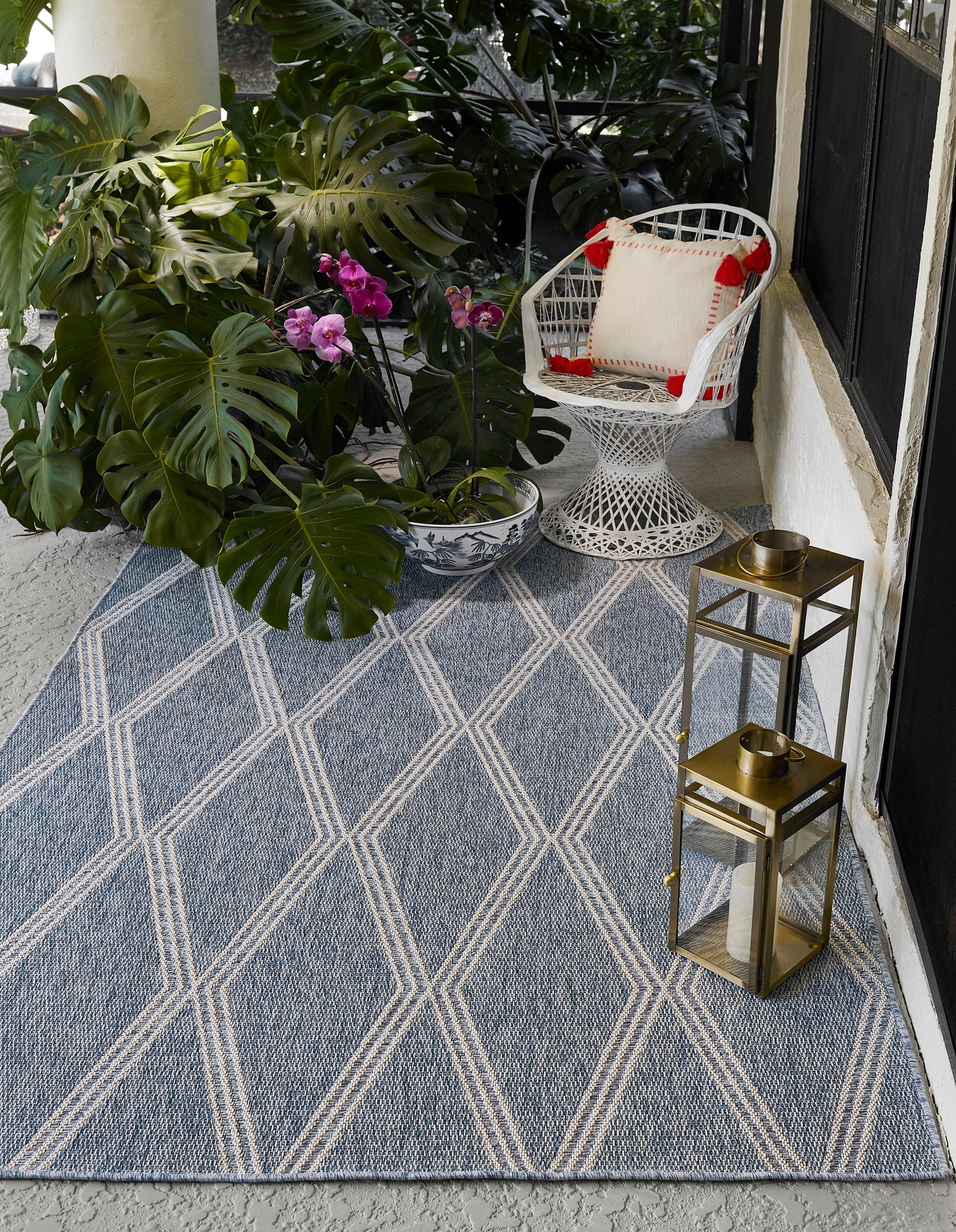 Blue geometric rug on a porch with monstera leaves, orchid pot, white wicker chair, and brass lanterns.