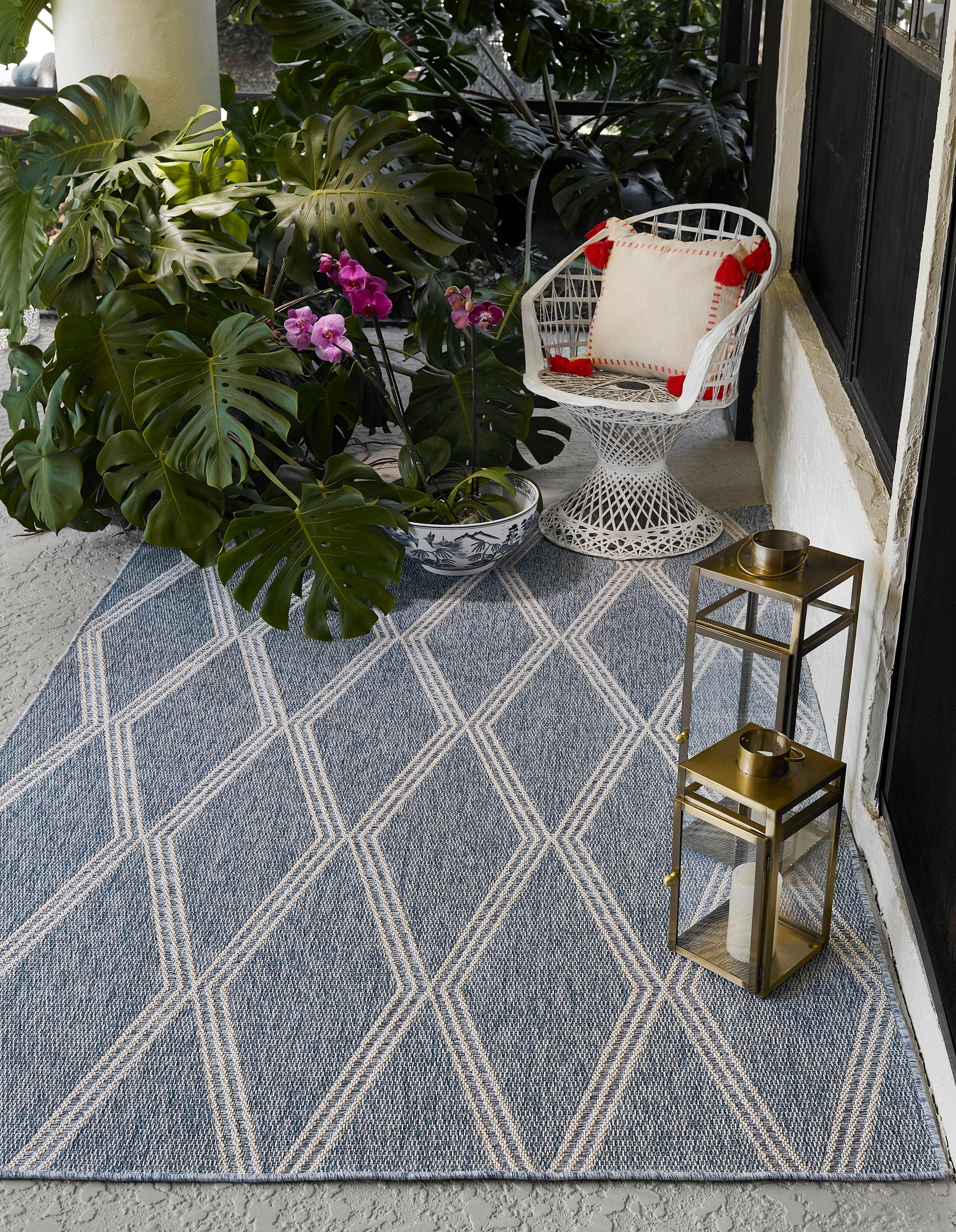 Blue geometric rug on a porch with monstera leaves, orchid pot, white wicker chair, and brass lanterns.