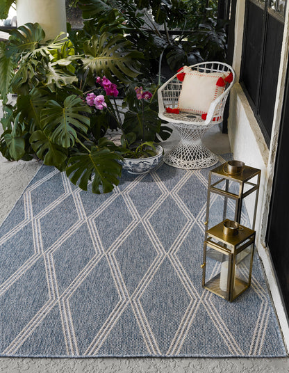Blue geometric rug on a porch with monstera leaves, orchid pot, white wicker chair, and brass lanterns.