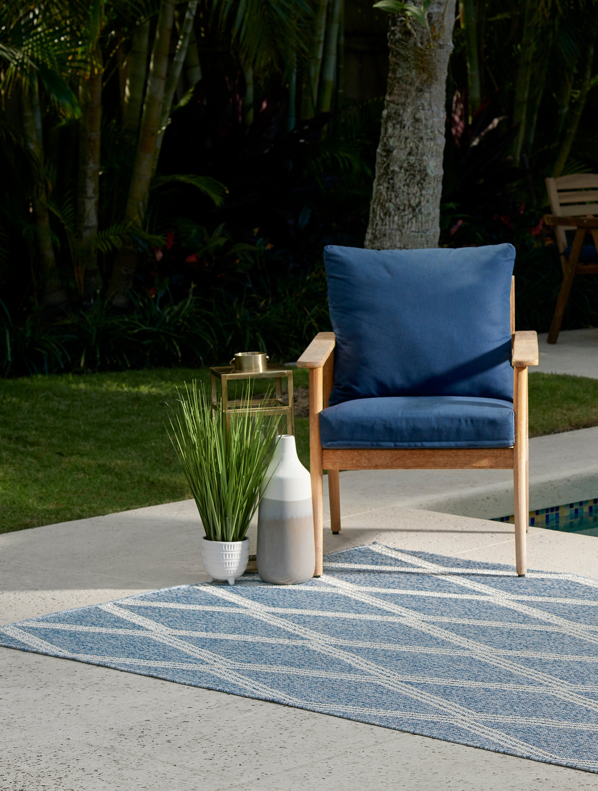 Wood outdoor armchair with navy cushions beside a blue geometric outdoor rug; gold metal side table, ceramic vases and potted grasses.