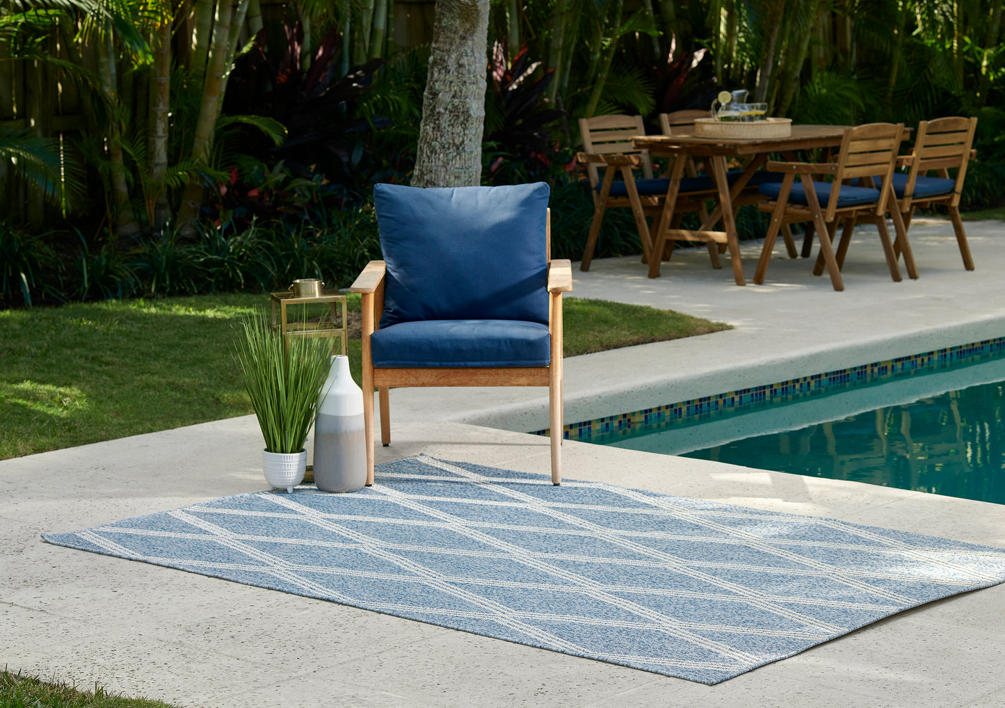 Blue cushioned wooden patio chair on a striped blue rug beside a pool, with plant, lantern & dining set in background.