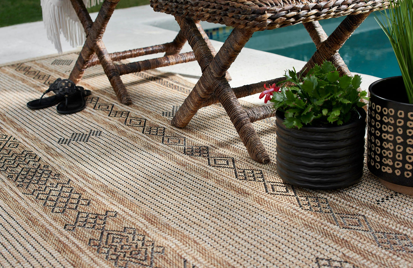 Natural fiber jute rug with black geometric patterns under a wicker chair, with black sandals and potted greenery by a pool.