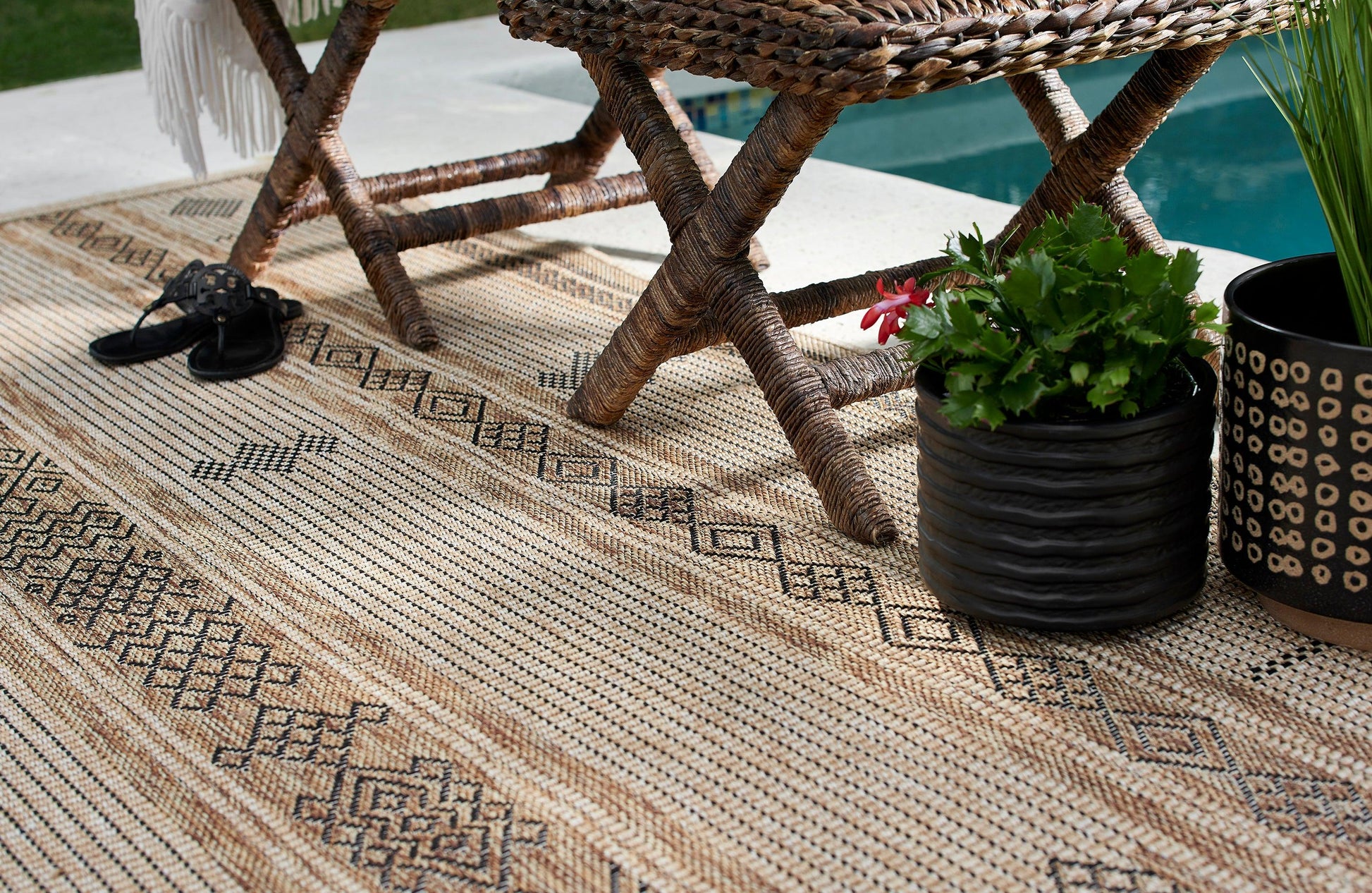 Natural fiber jute rug with black geometric patterns under a wicker chair, with black sandals and potted greenery by a pool.