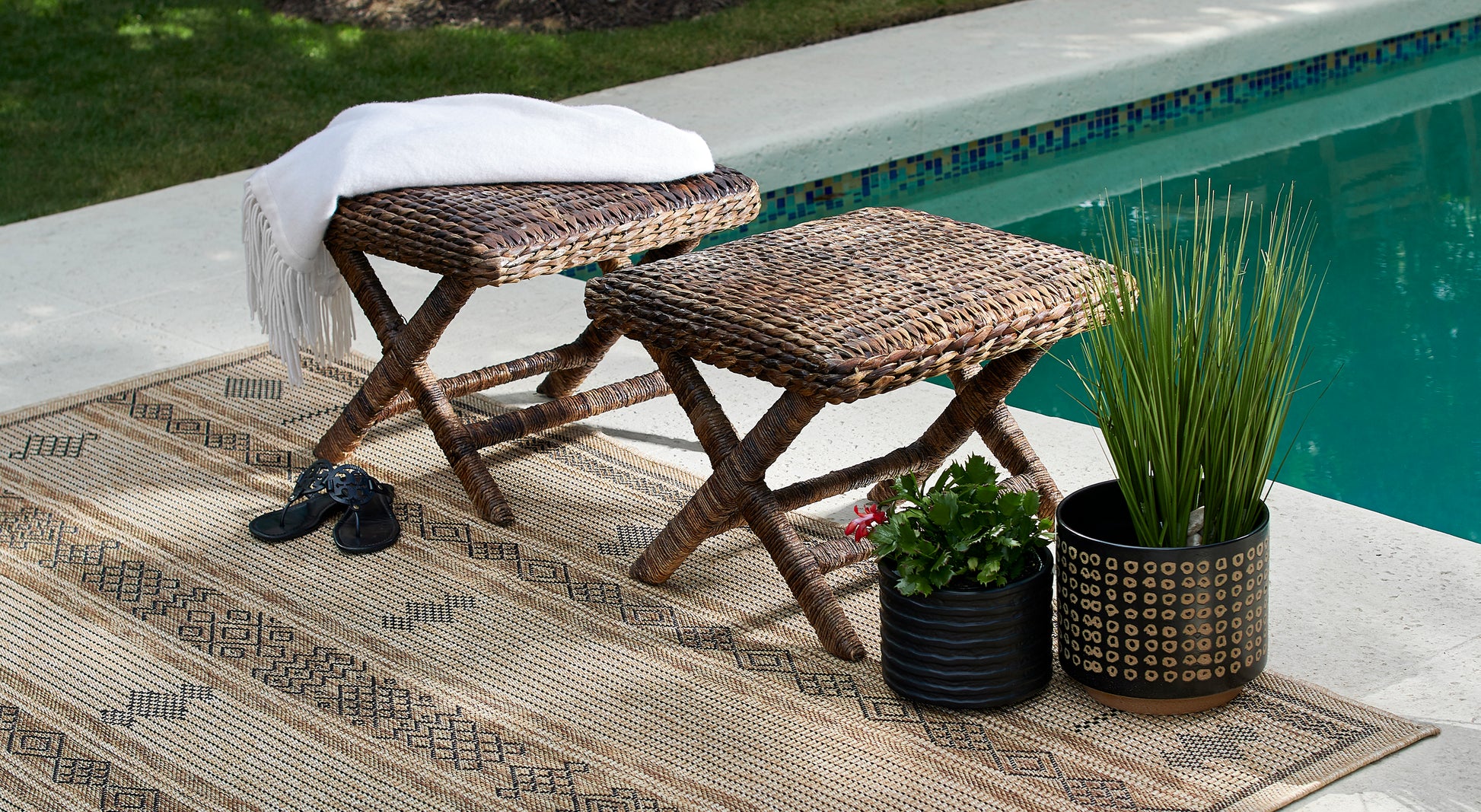 Natural wicker stools with a white fringe throw, beige geometric rug, sandals, and two planters by a turquoise pool.