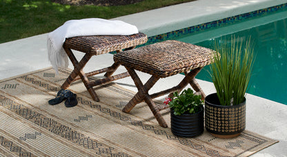 Natural wicker stools with a white fringe throw, beige geometric rug, sandals, and two planters by a turquoise pool.