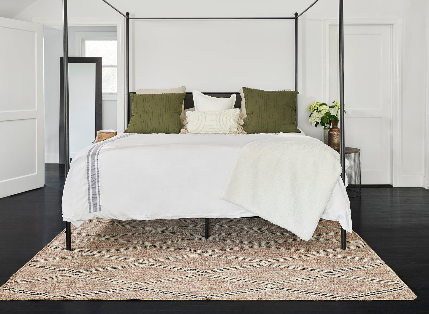 Black metal canopy bed with white bedding, olive green pillows & cream throw, on a woven natural rug.