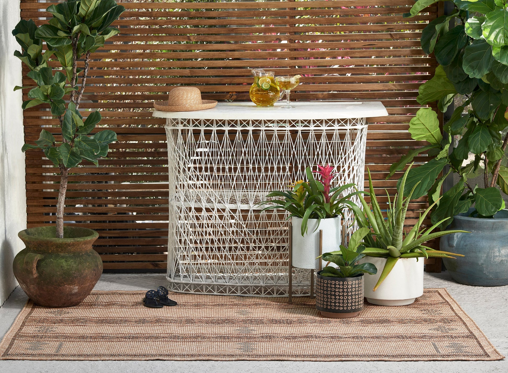 Outdoor patio with white woven lattice bar cabinet, straw hat, pitcher and glasses, wood slat screen, potted plants on rug.