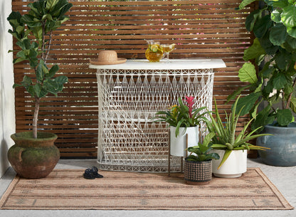 Outdoor patio with white woven lattice bar cabinet, straw hat, pitcher and glasses, wood slat screen, potted plants on rug.