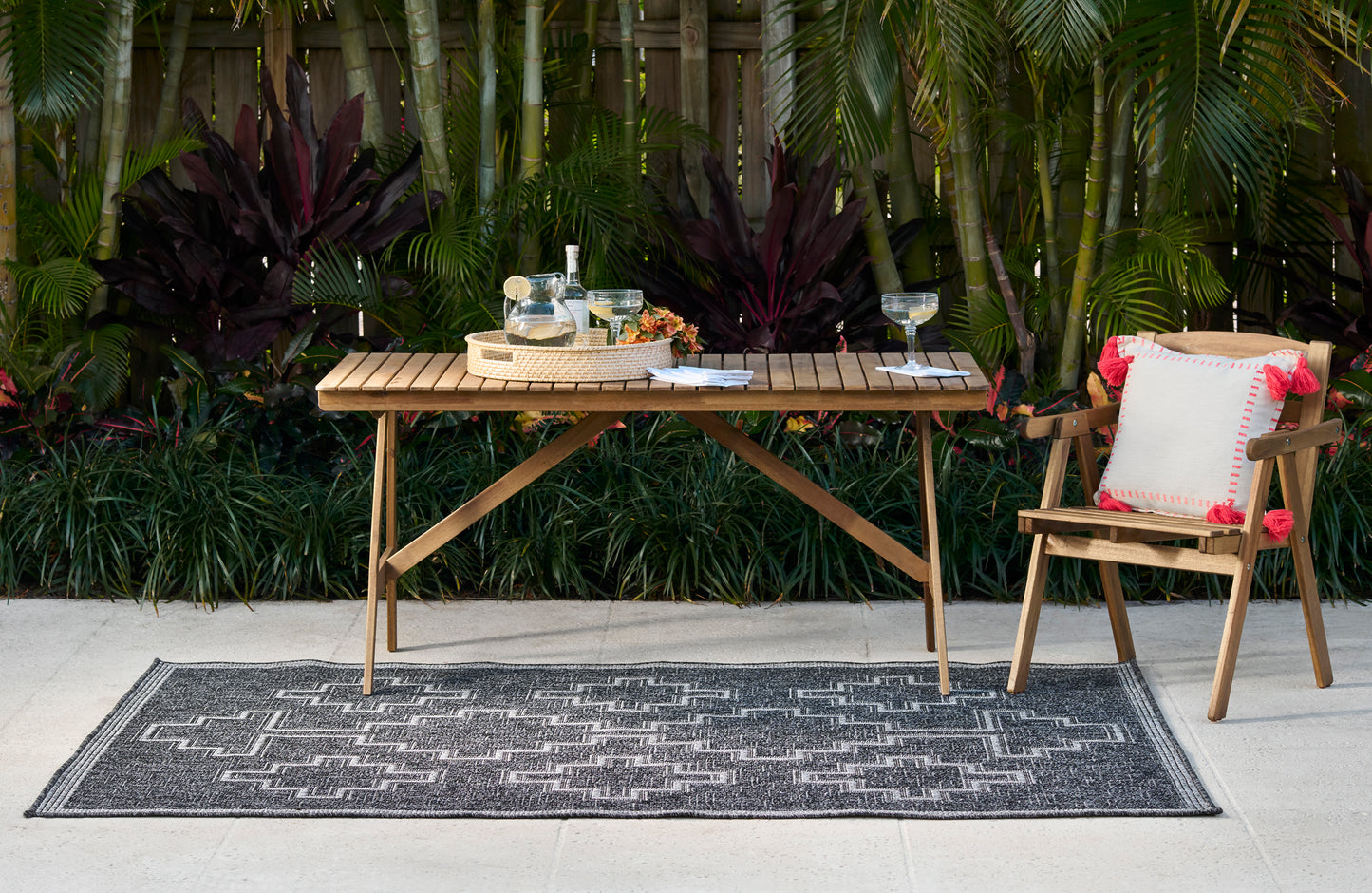 Wooden outdoor dining set on a black geometric rug, slatted table and simple chair with white cushion, tropical backdrop