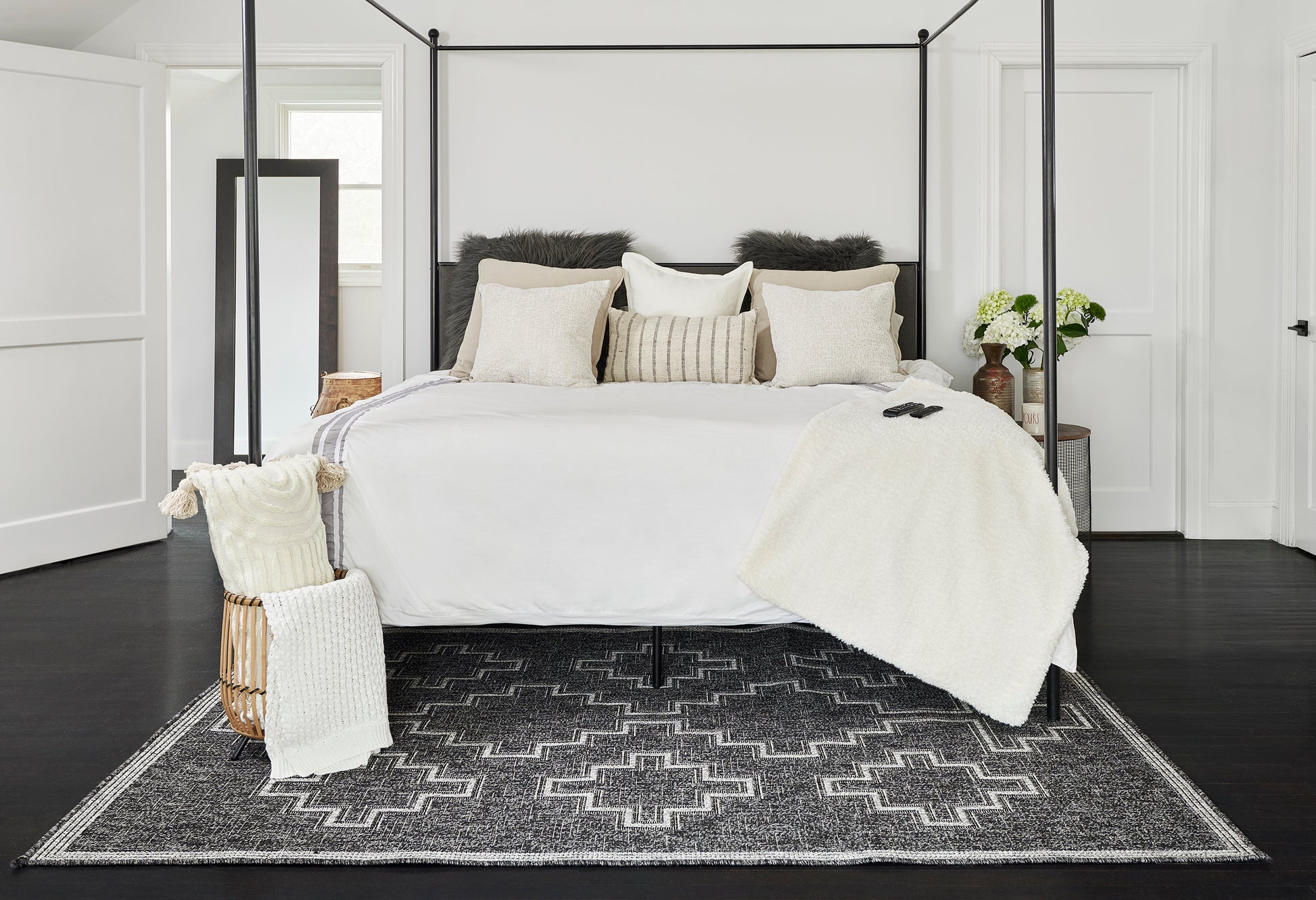 White four-poster bed with black metal frame, cream bedding, beige and faux-fur pillows, white throw, gray geometric rug.