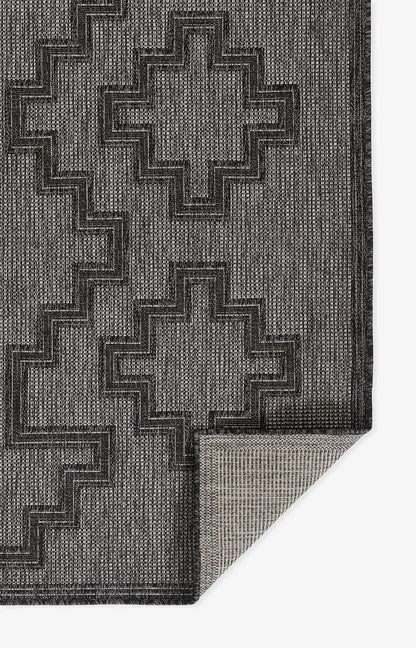 Dark gray geometric rug with stepped cross motif, textured weave; folded corner shows lighter underside.