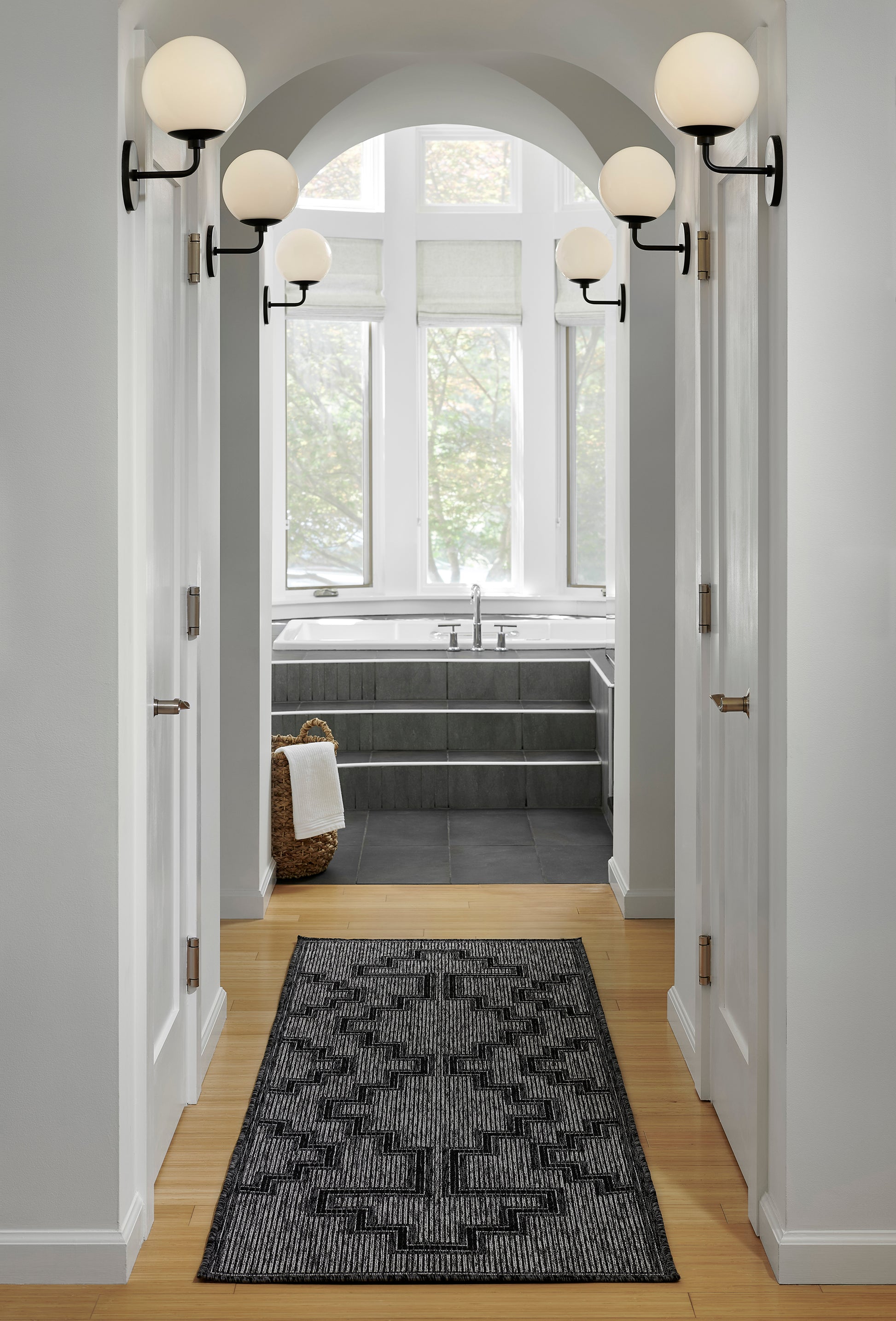 Hallway to a spa bath with arched ceiling, globe sconces, dark tile steps to a tub, and a charcoal geometric rug.