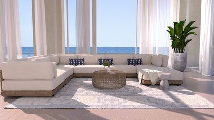 Blue 8' x 10' rug in a coastal living room with a beige sectional, navy patterned cushions, a round wicker coffee table, throw blanket, potted plant, and an ocean view.