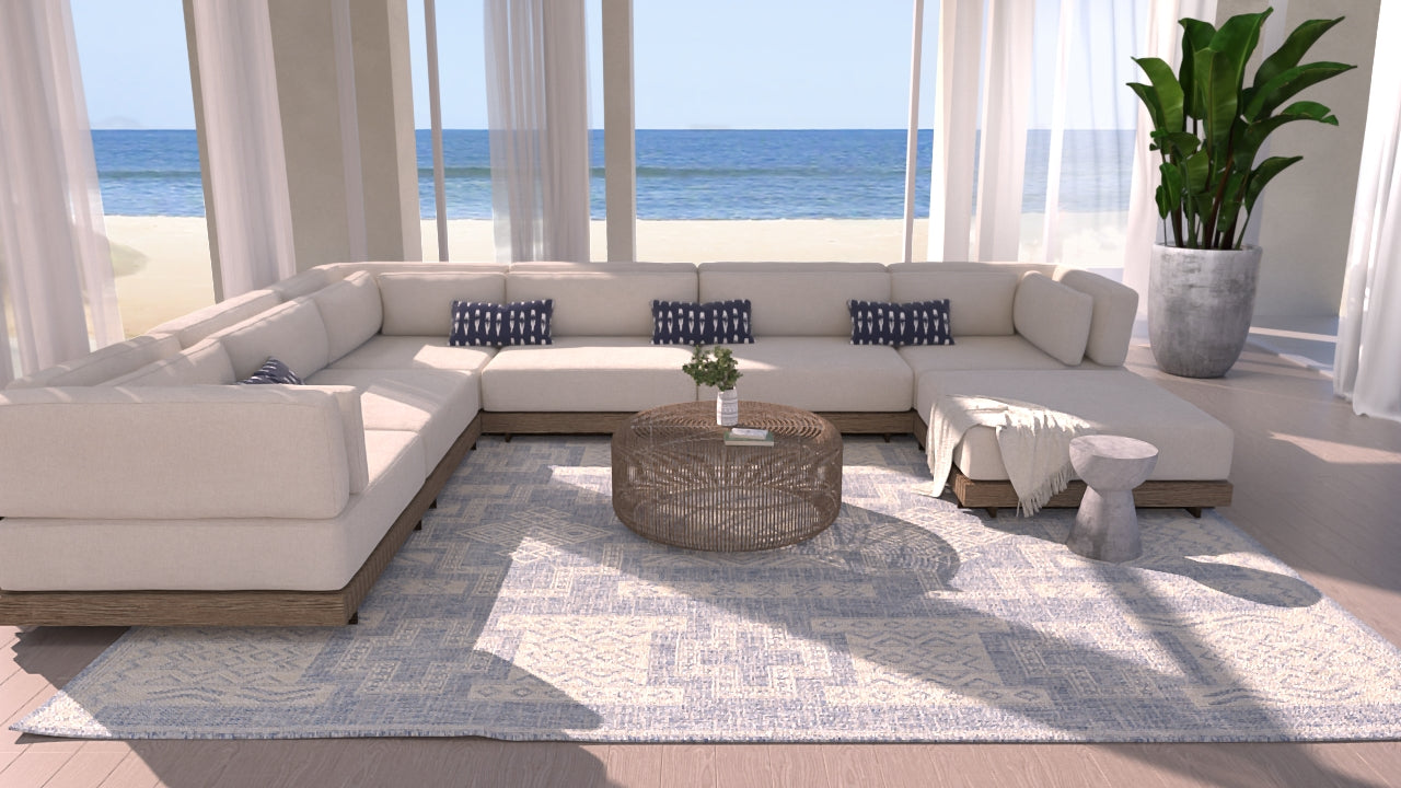 Coastal living room with cream sectional, round wicker coffee table, blue cushions, rug, plant, curtains, ocean view